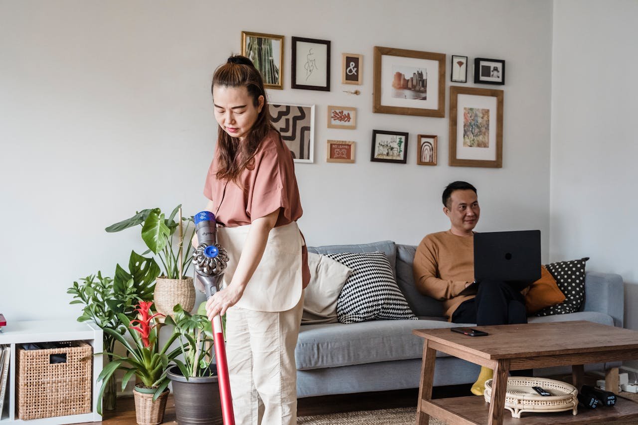 An Asian woman vacuuming while a man works on a laptop in a modern living room.